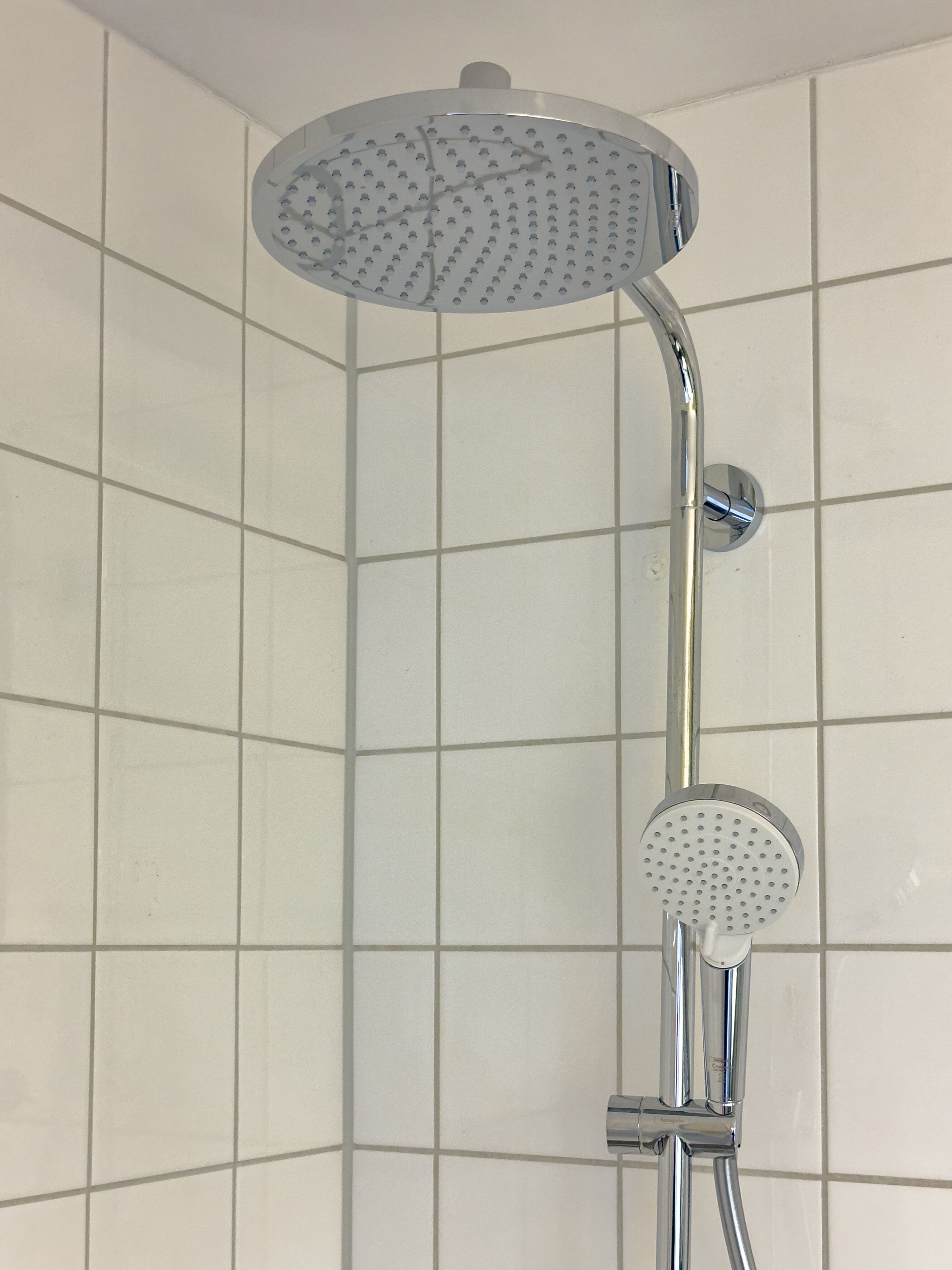 Ducha de esquina con cabezal redondo y un cabezal de ducha manual más pequeño, montado en un accesorio de cromo sobre una pared con azulejos.