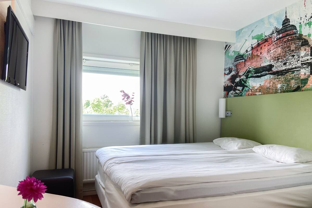 A hotel room with a bed, a window with curtains, a painting on the wall, and a vase with flowers on a table.