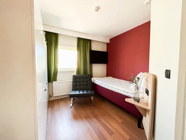 A bedroom with a bed, a chair, a radiator, a window with curtains, and a wall-mounted TV.