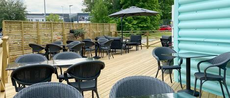 An outdoor patio with tables and chairs, a wooden deck, and a blue building in the background.