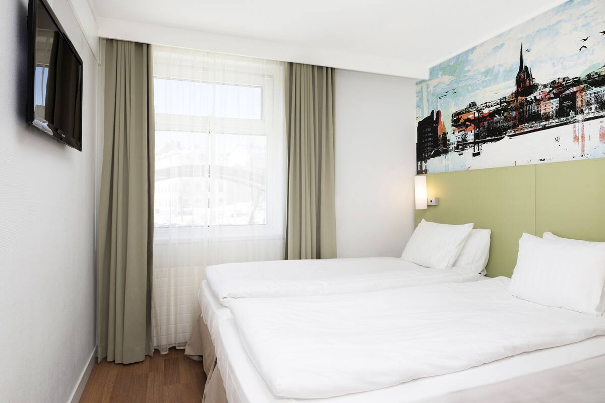 Una habitación de hotel con dos camas, un ventanal con cortinas translúcidas, un televisor de pantalla plana en la pared y un cuadro con un paisaje urbano.