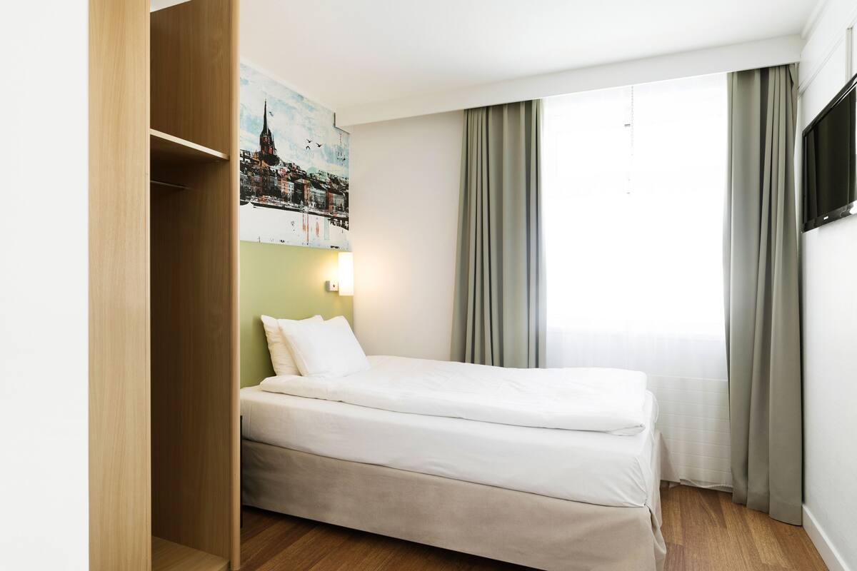 Habitación de hotel con cama, armario, televisor montado en la pared y ventana con cortinas.