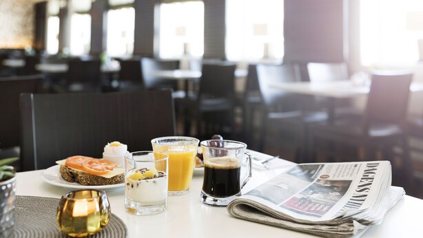 Un desayuno con un periódico, un vaso de zumo de naranja, un café y un plato con un sándwich y un postre.