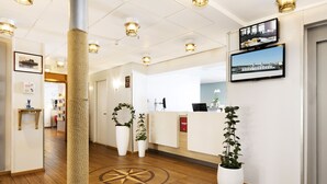 Zona de recepción de hotel con suelo de madera, mostrador, plantas en macetas y televisor en la pared.
