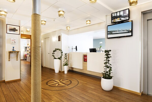 A hotel reception area with a wooden floor, a reception desk, potted plants, and a wall-mounted television.