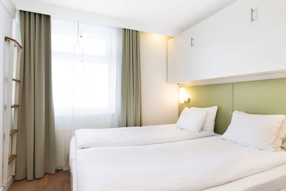 Habitación de hotel con dos camas, una ventana con cortinas, una lámpara de pared y cabecera verde.