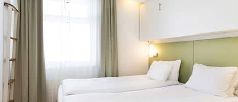 Une chambre d’hôtel avec deux lits, une fenêtre avec des rideaux, un lampadaire fixé au mur et une tête de lit verte.