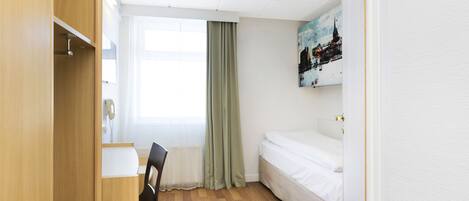 Quarto pequeno e bem iluminado, com uma escrivaninha de madeira, uma cadeira, uma cama e uma janela com cortinas.
