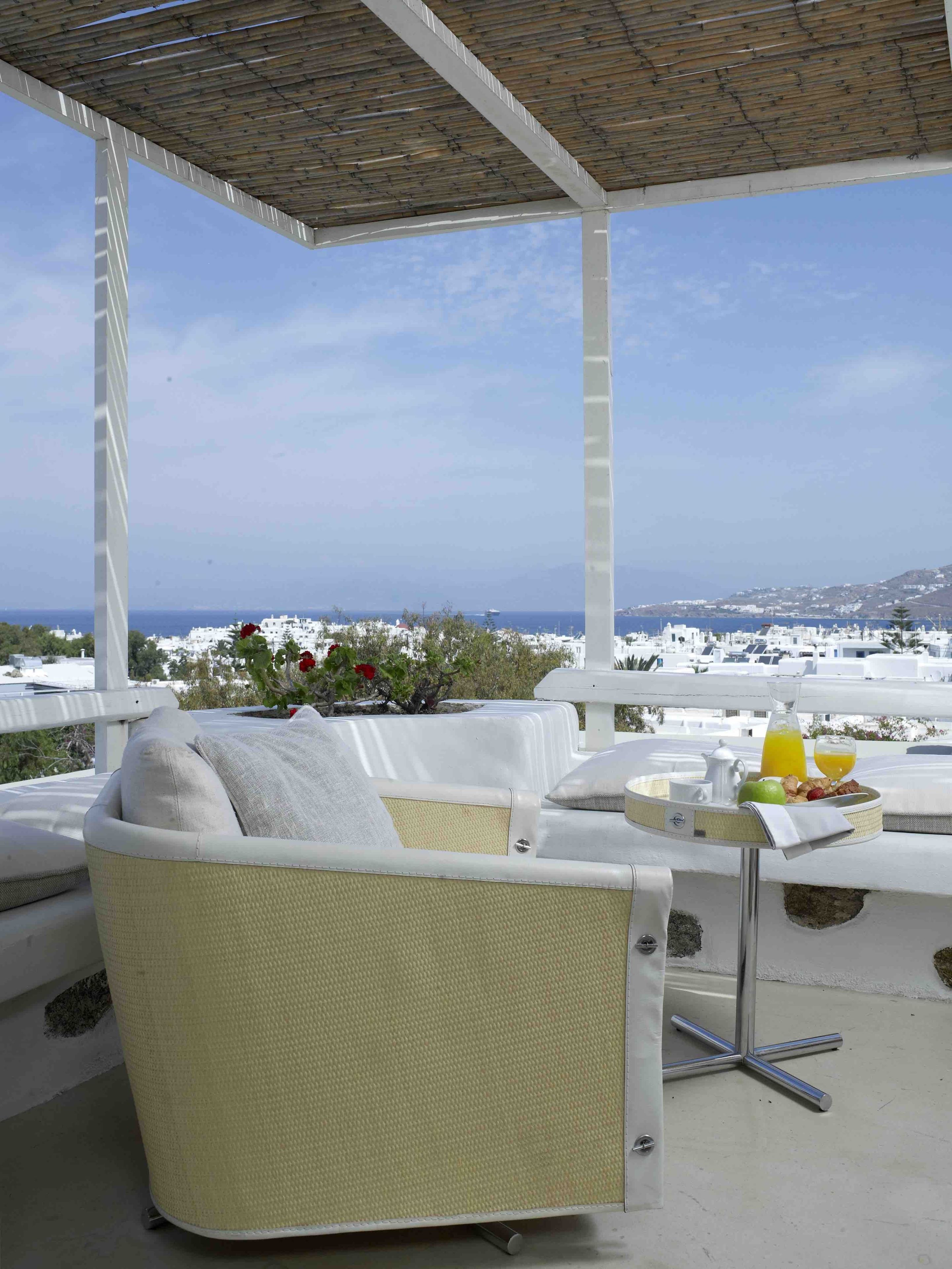 A covered outdoor seating area with a view of the sea and a white building in the background.