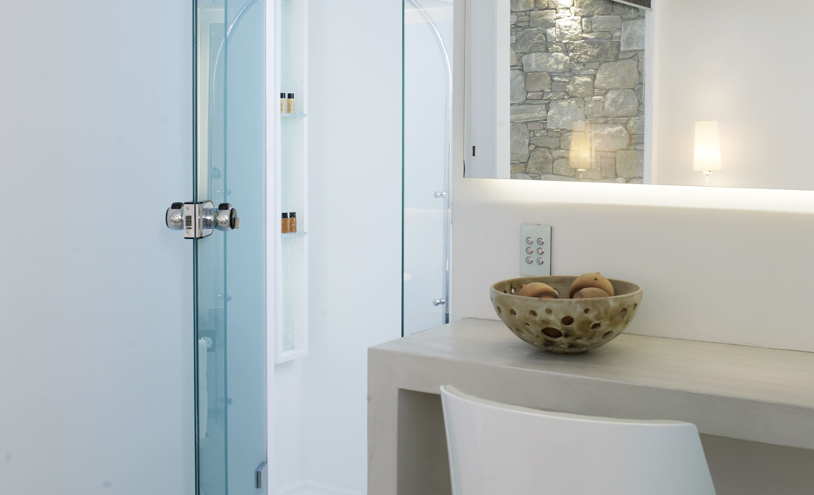 A bathroom with a glass shower door, a stone wall, a bowl of toiletries on a counter, and a white chair.