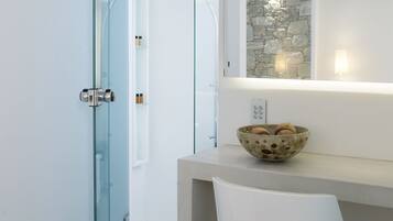 A bathroom with a glass shower door, a stone wall, a bowl of toiletries on a counter, and a white chair.