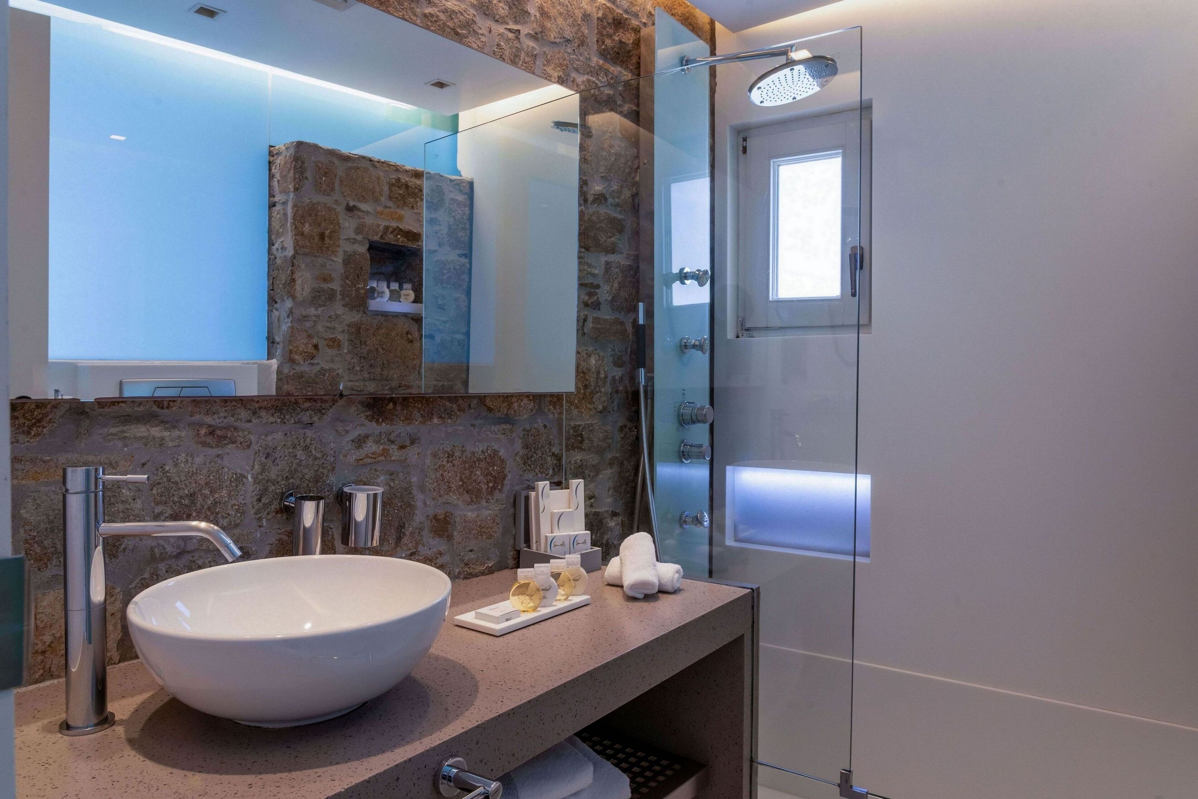 A modern bathroom with a stone wall, a large white sink, a glass shower enclosure, and a mirror above the sink.