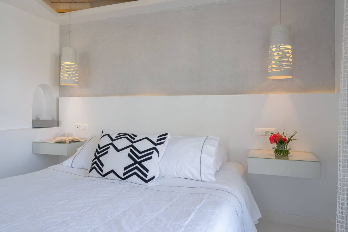 Un dormitorio moderno con cama, ropa de cama blanca, almohada estampada, mesitas de noche con lámparas y un florero con flores.
