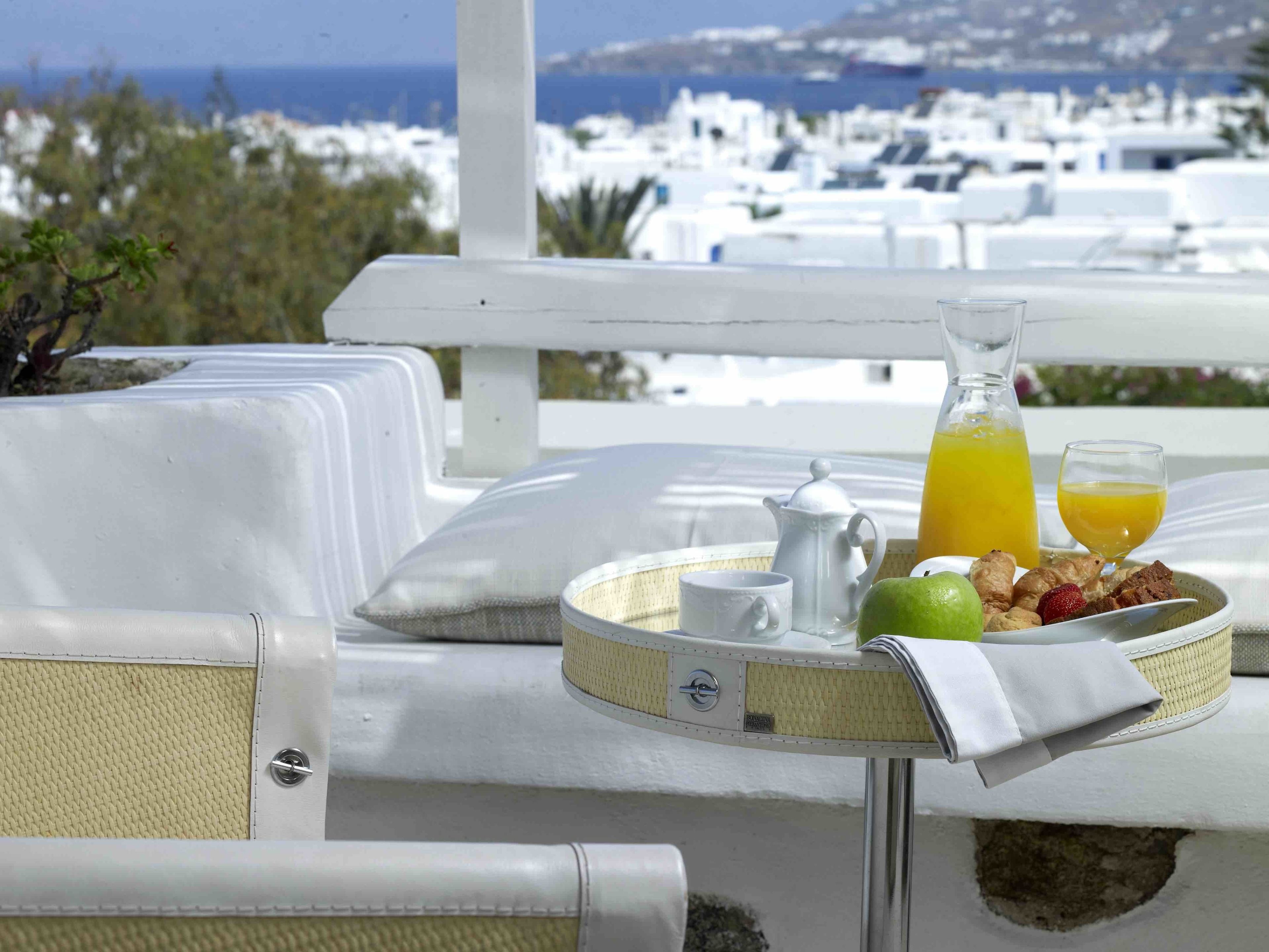 A breakfast setup with a pitcher of orange juice, a cup, a plate with fruits and pastries, and a teapot on a balcony overlooking a coastal town.
