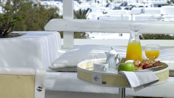 A breakfast setup with a pitcher of orange juice, a cup, a plate with fruits and pastries, and a teapot on a balcony overlooking a coastal town.