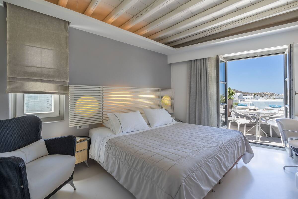 Un dormitorio moderno con una cama grande, una mesita de noche, una silla y un balcón con mobiliario exterior.