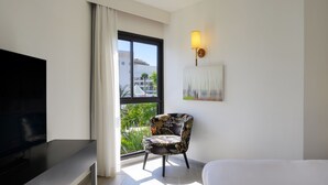 A hotel room with a bed, a TV, a chair, a painting, and a window with a view of buildings and greenery. - Riviera by Isrotel Collection (Eilat)