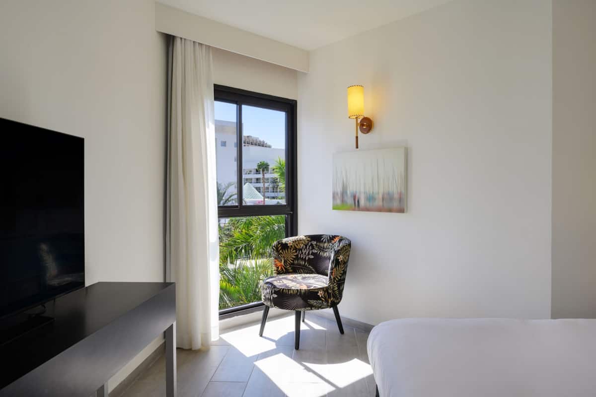 A hotel room with a bed, a TV, a chair, a painting, and a window with a view of buildings and greenery.
