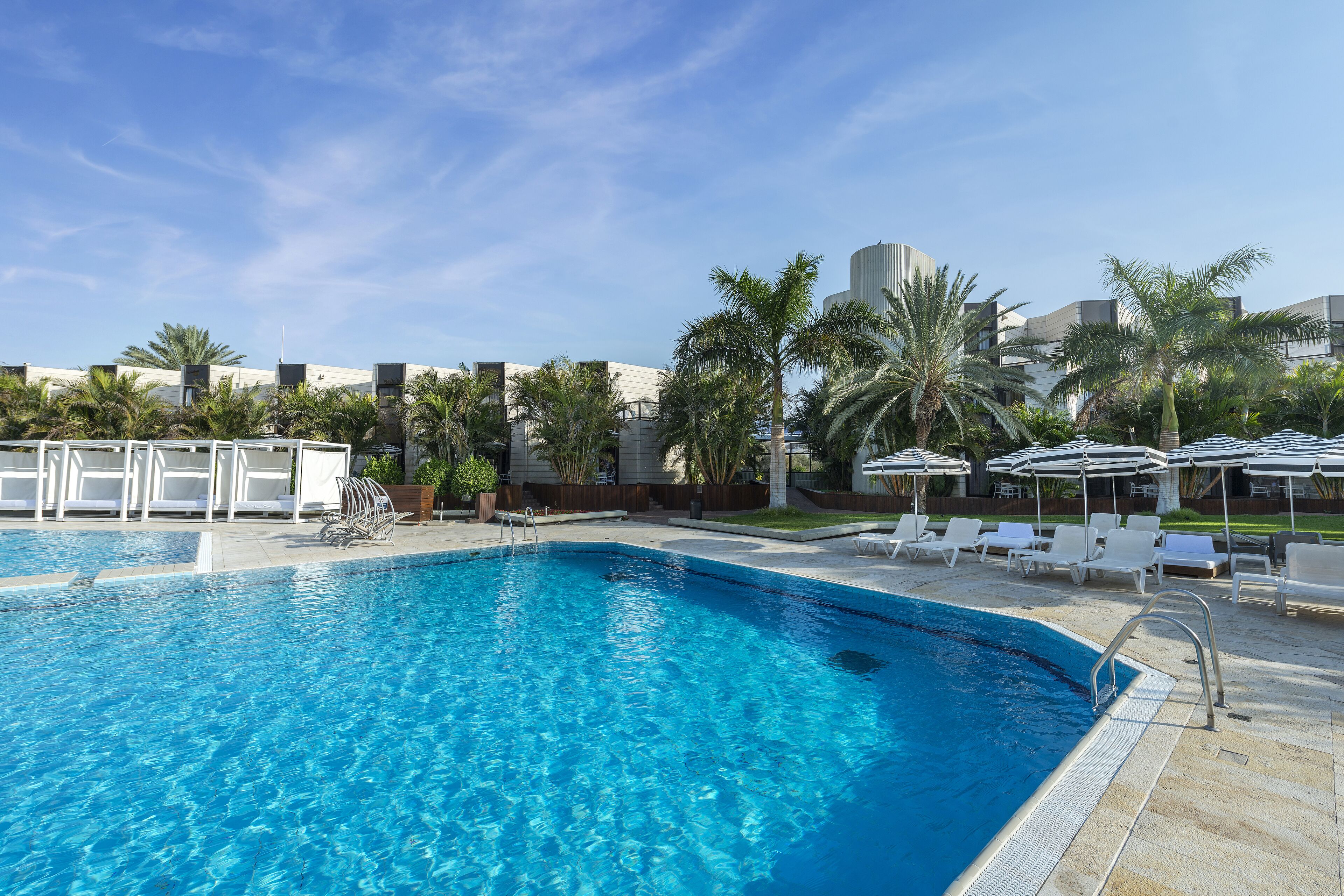 A swimming pool with lounge chairs and umbrellas, surrounded by palm trees and modern buildings.
