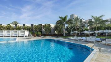 A swimming pool with lounge chairs and umbrellas, surrounded by palm trees and modern buildings.