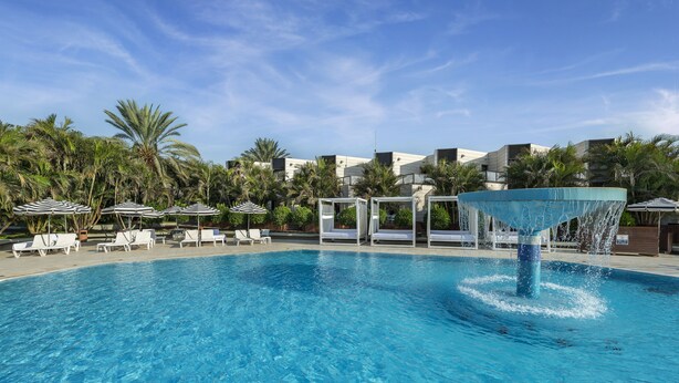 A swimming pool with lounge chairs and umbrellas, surrounded by palm trees and modern buildings. - Riviera by Isrotel Collection (Eilat)