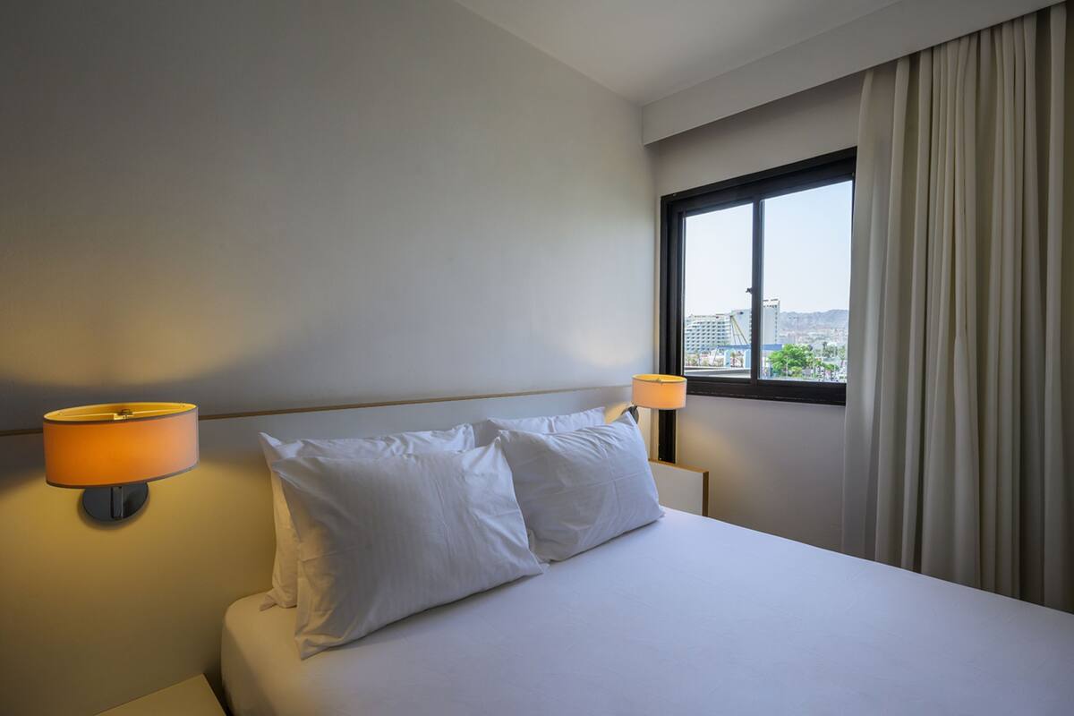 A hotel room with a bed, two pillows, a nightstand, a lamp, a window with curtains, and a view of buildings outside.