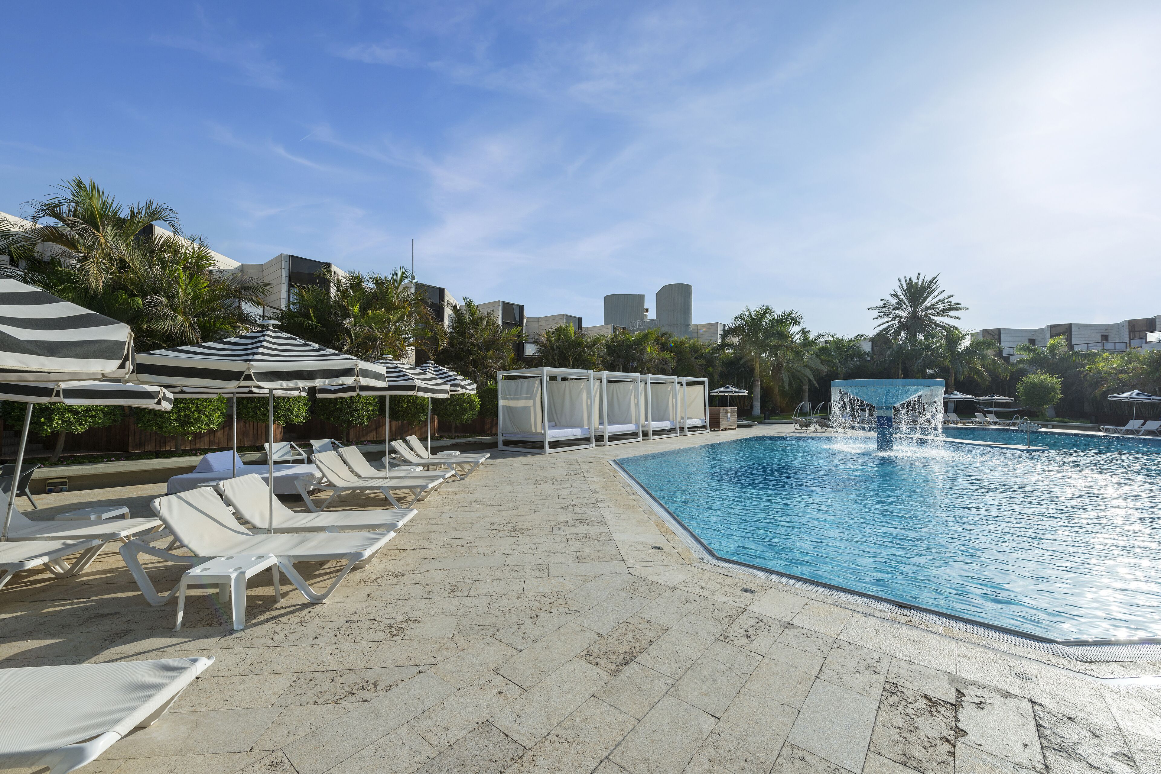 A swimming pool area with lounge chairs and umbrellas, surrounded by palm trees and modern buildings.