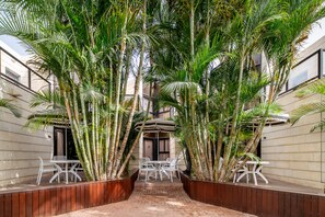 Un patio con un área central de estar rodeado de altas palmeras, un espacio cubierto para sentarse y un edificio con fachada de tejas.