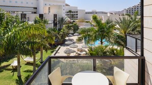A balcony with a glass railing, two white chairs, and a small round table. In the background, there is a swimming pool surrounded by palm trees and lounge chairs. - Riviera by Isrotel Collection (Eilat)