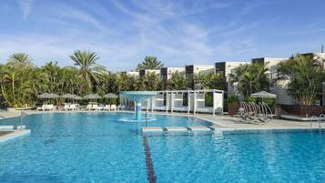 A swimming pool with lounge chairs and umbrellas, surrounded by palm trees and modern buildings.