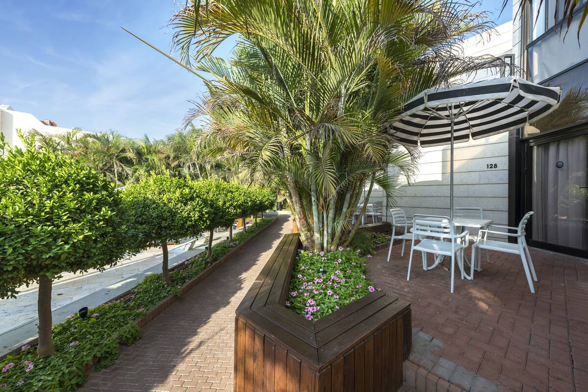 A paved walkway lined with lush greenery and a wooden planter with pink flowers.