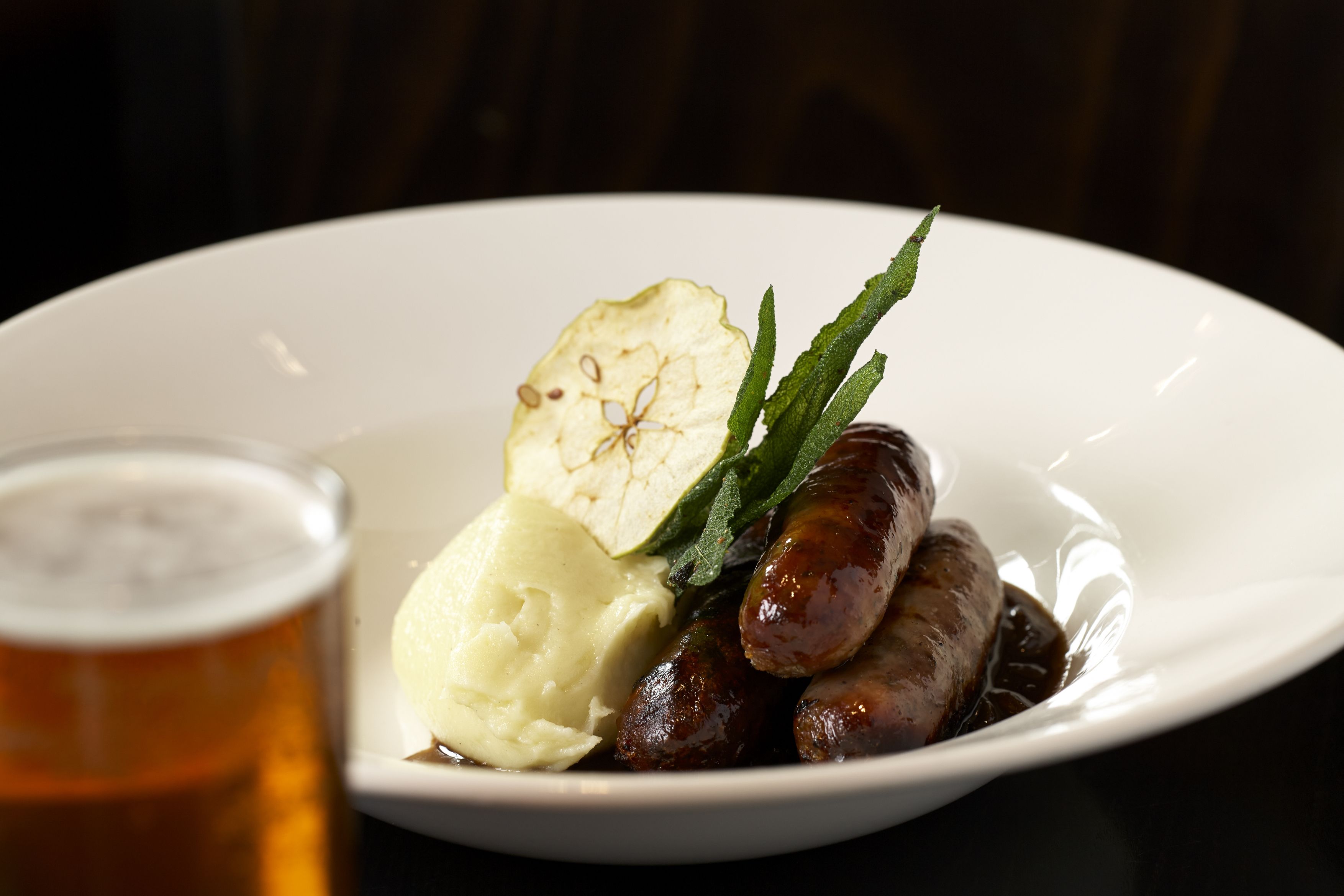 A plate with sausages, mashed potatoes, and a slice of apple, accompanied by a glass of beer.