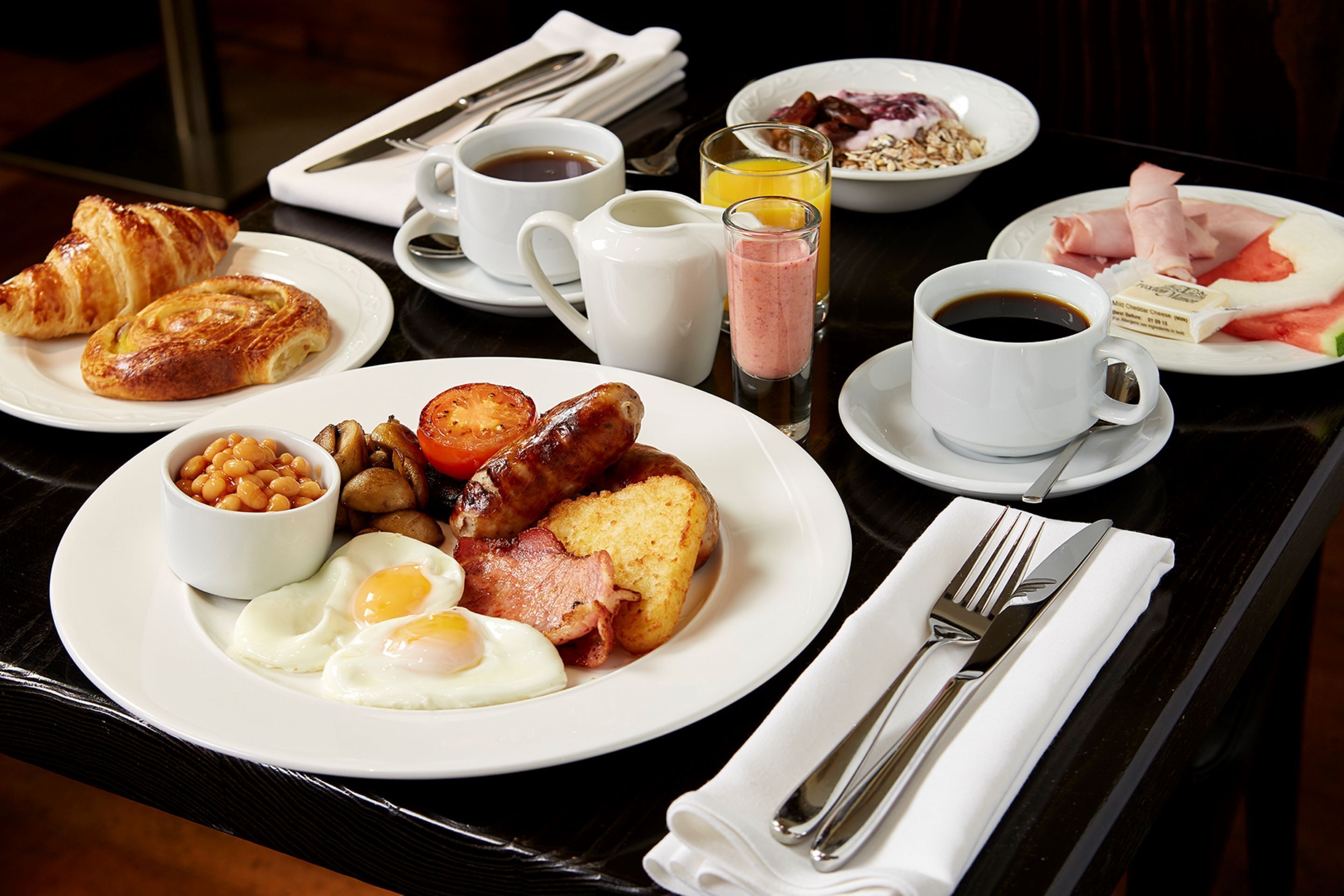 Una mesa de desayuno con platos de huevos, salchichas, tostadas y frijoles. También hay tazas de café y jugo.