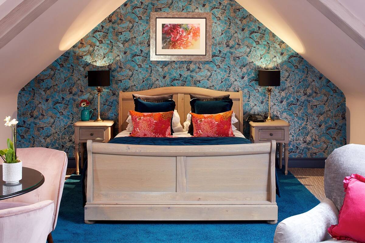 A bedroom with a high ceiling, a wooden bed, two bedside tables with lamps, a framed picture on the wall, and a blue carpet.