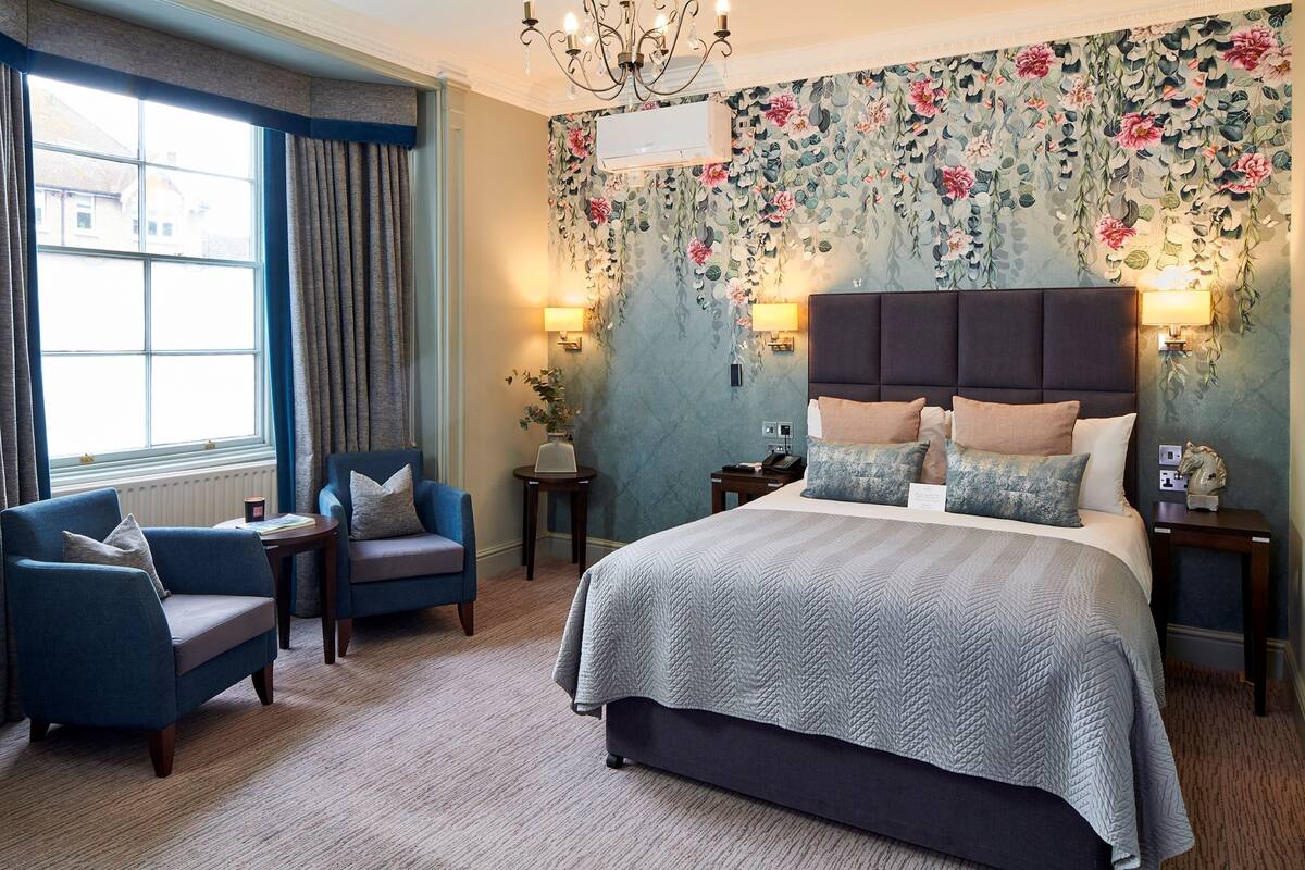 A bedroom with a large bed, two armchairs, a chandelier, floral wallpaper, and a window with curtains.
