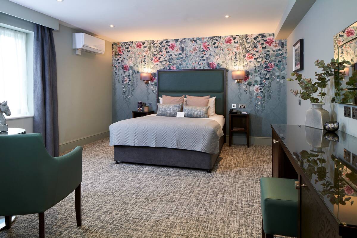 A modern bedroom with a large bed, two bedside tables, a green armchair, and a desk with a vase of flowers.