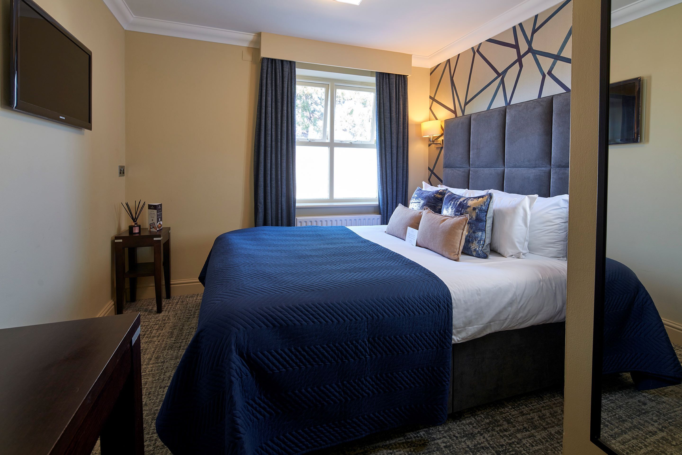 A hotel room with a large bed, a bedside table, a window with curtains, and a wall-mounted TV.