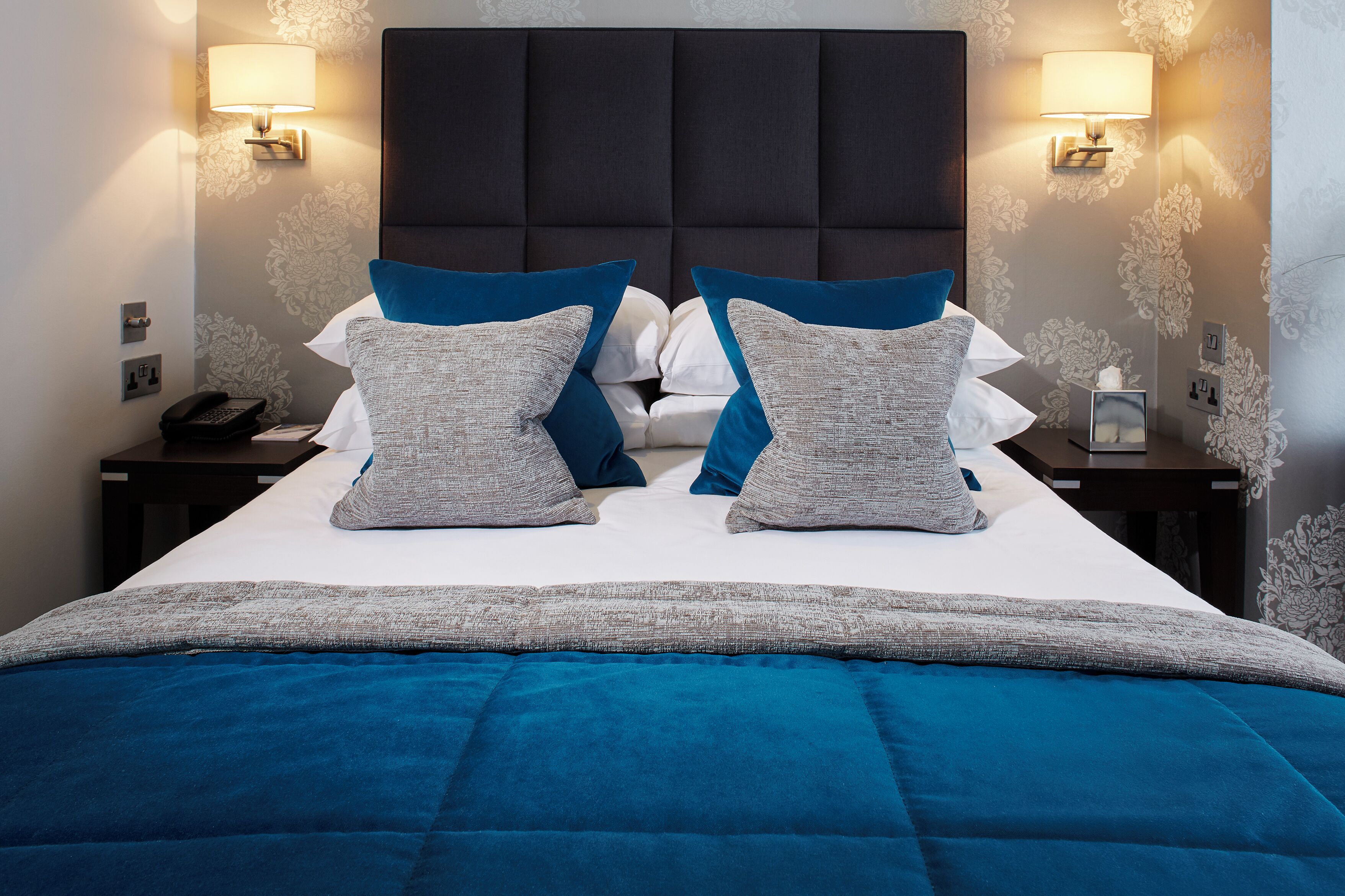 Una cama bien hecha con edredón azul y gris, dos lámparas de noche y una mesita de noche con un teléfono y una pequeña mesa.