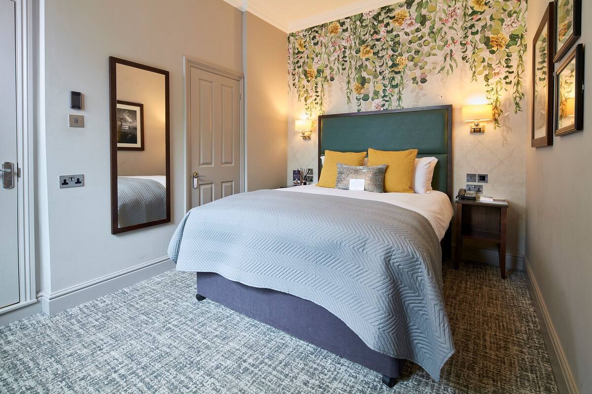 A neatly arranged bedroom with a large bed, a bedside table, a mirror, and a wall-mounted lamp.