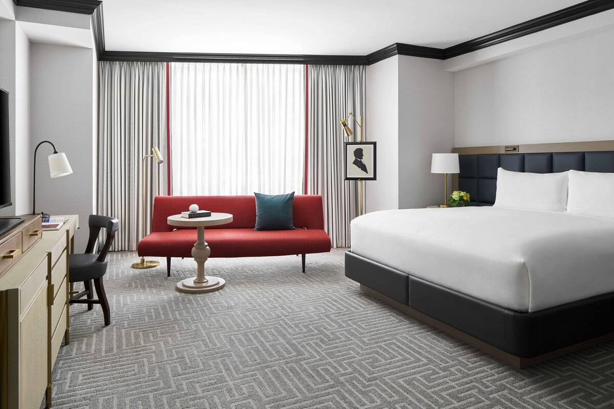 A hotel room with a large bed, a red sofa, a small round table, a desk with a chair, and a large window with curtains.