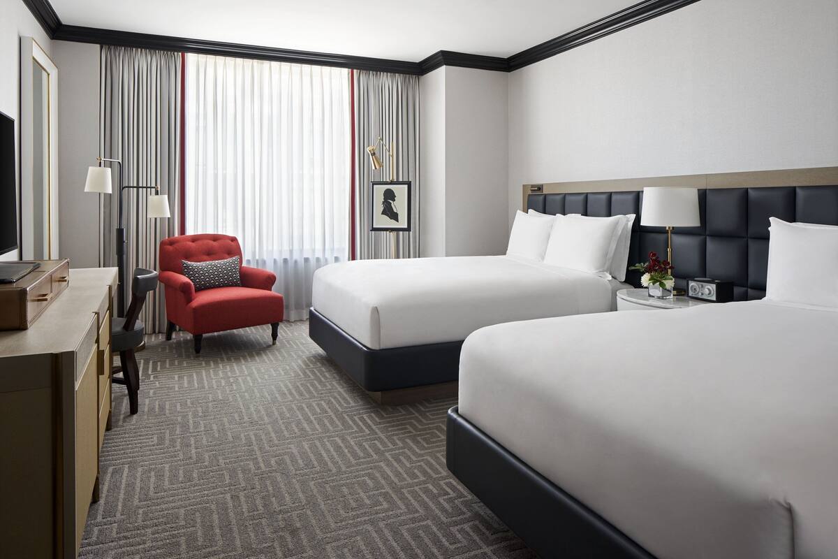 A hotel room with two beds, a desk, a red armchair, a large window with curtains, and a framed picture on the wall.