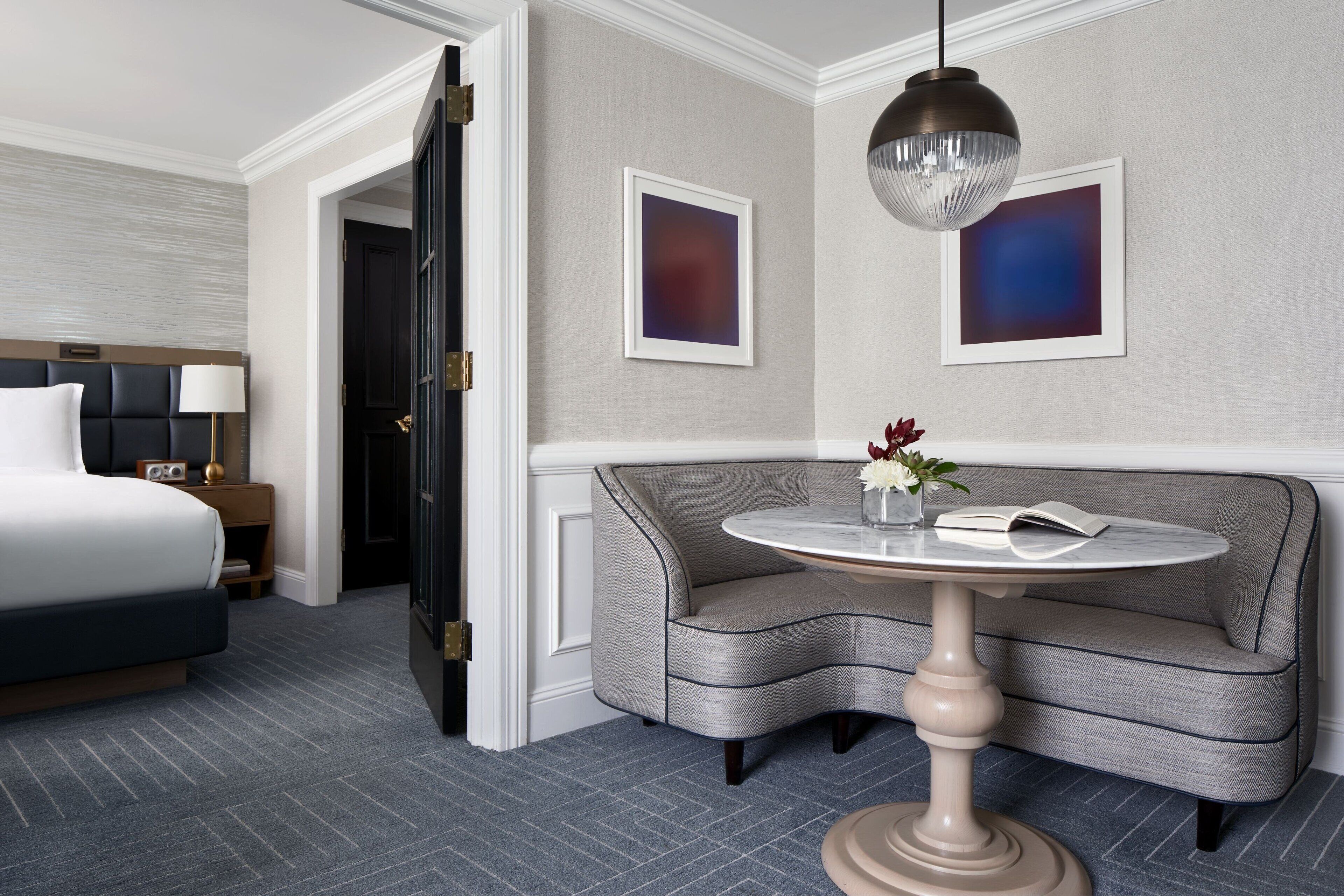 A hotel room with a bed, a sofa, a round table with a vase and book, and two framed pictures on the wall.