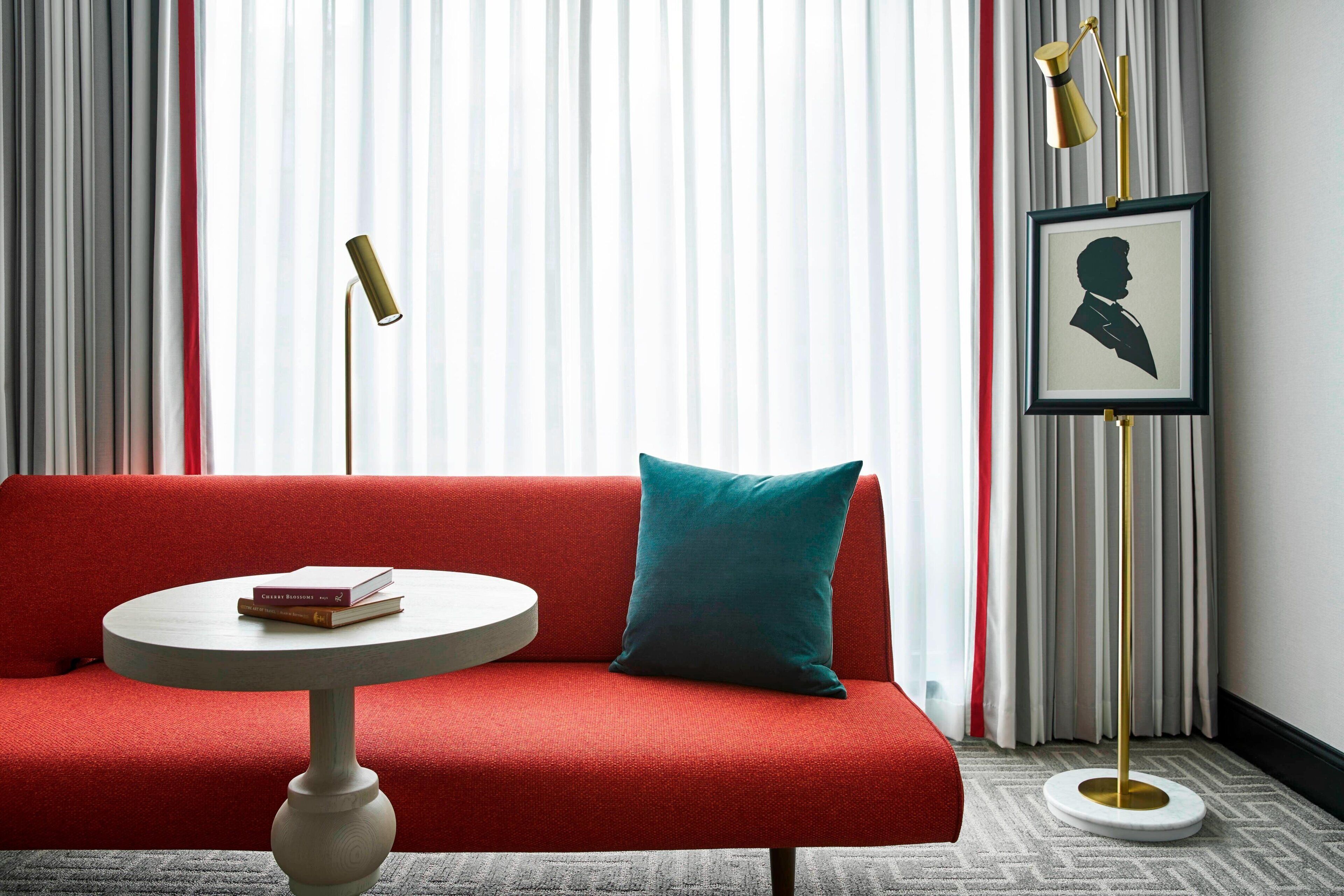 A red sofa with a teal pillow, a round white table with books, and a framed silhouette portrait on a gold floor lamp.