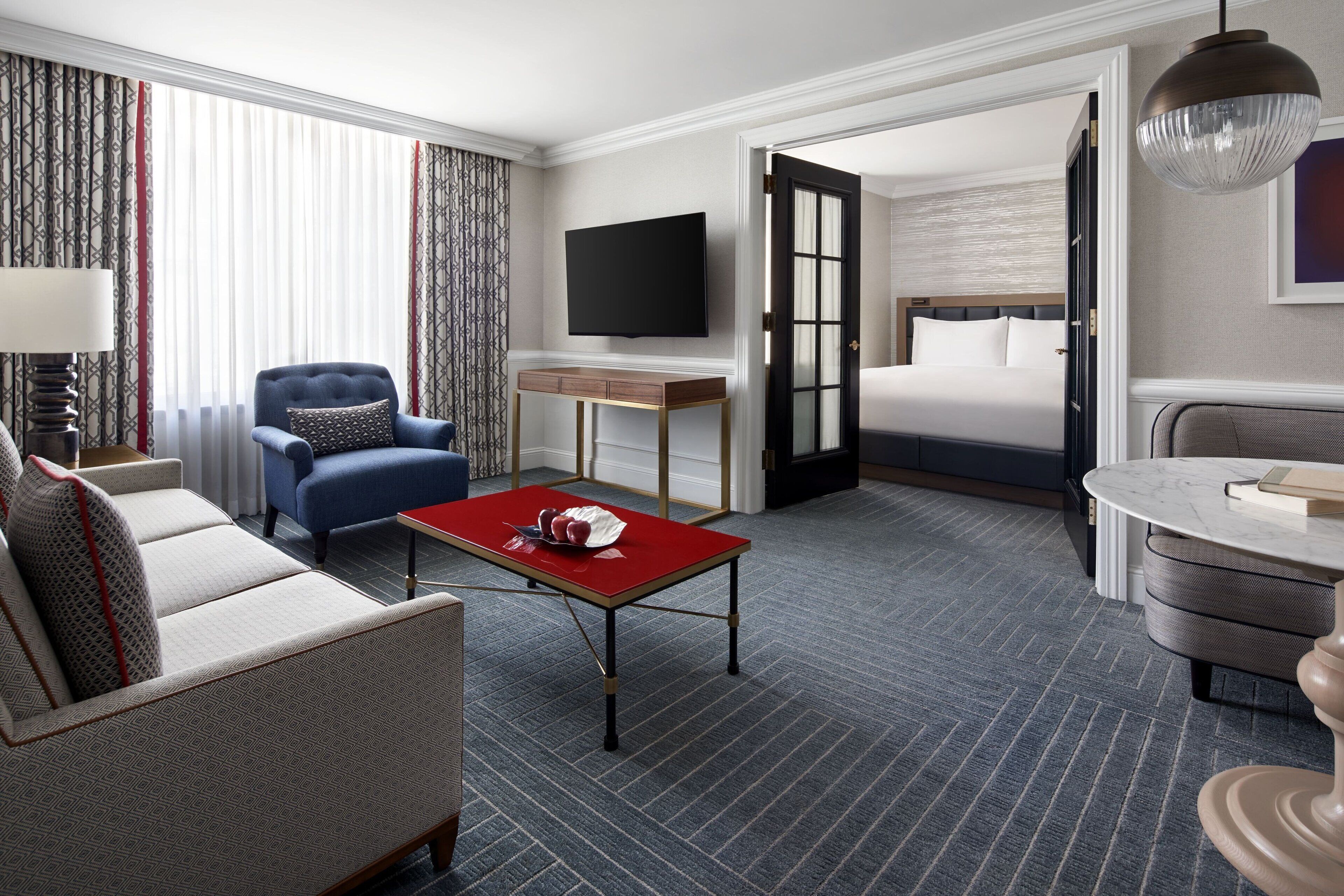 A hotel room with a sofa, armchair, a small table with a red top, a TV mounted on the wall, and a bed with a wooden headboard.