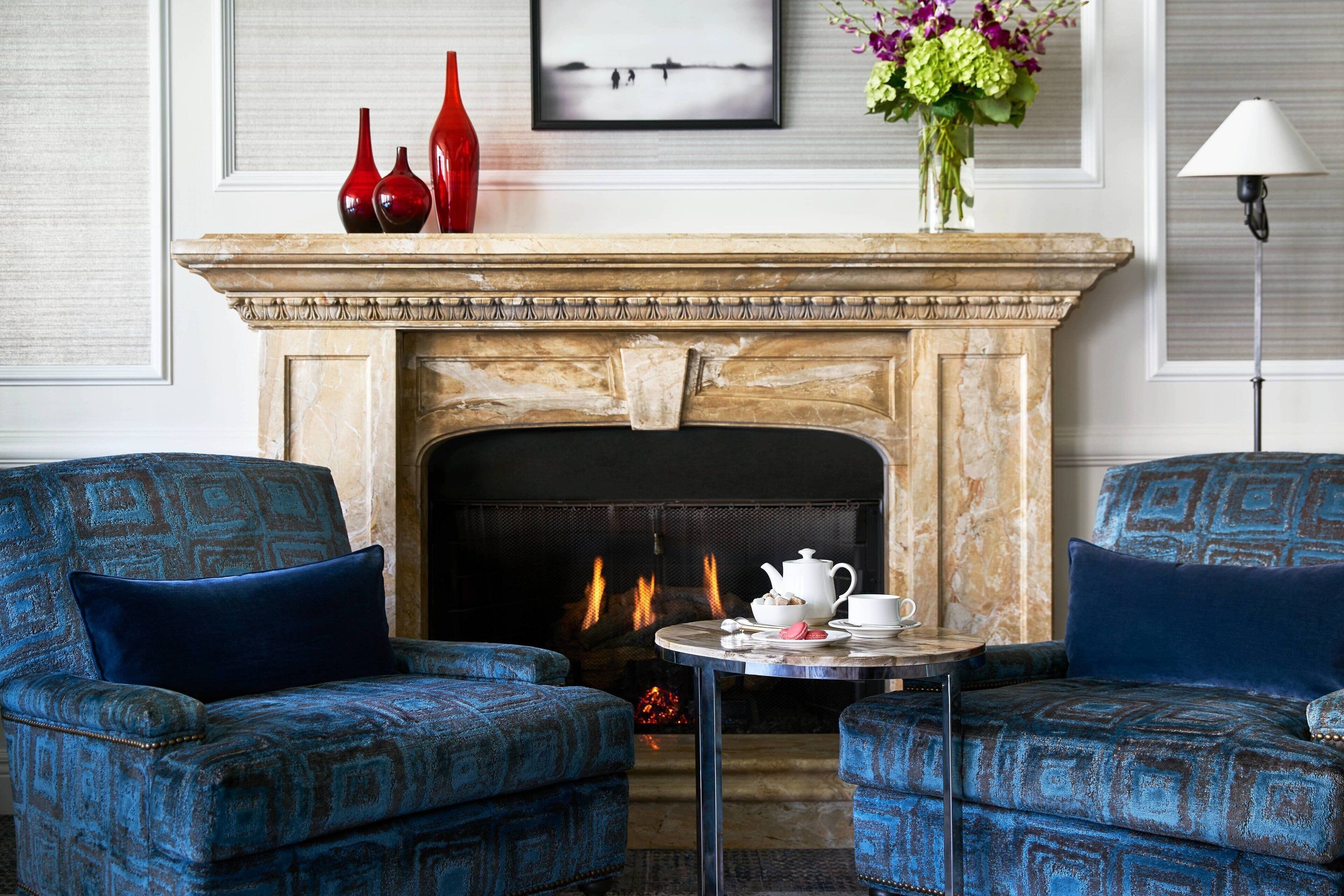 A room with a fireplace, two patterned armchairs, a small table with a teapot and cups, and a vase with flowers.