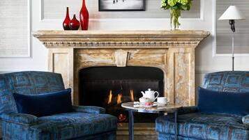 A room with a fireplace, two patterned armchairs, a small table with a teapot and cups, and a vase with flowers.