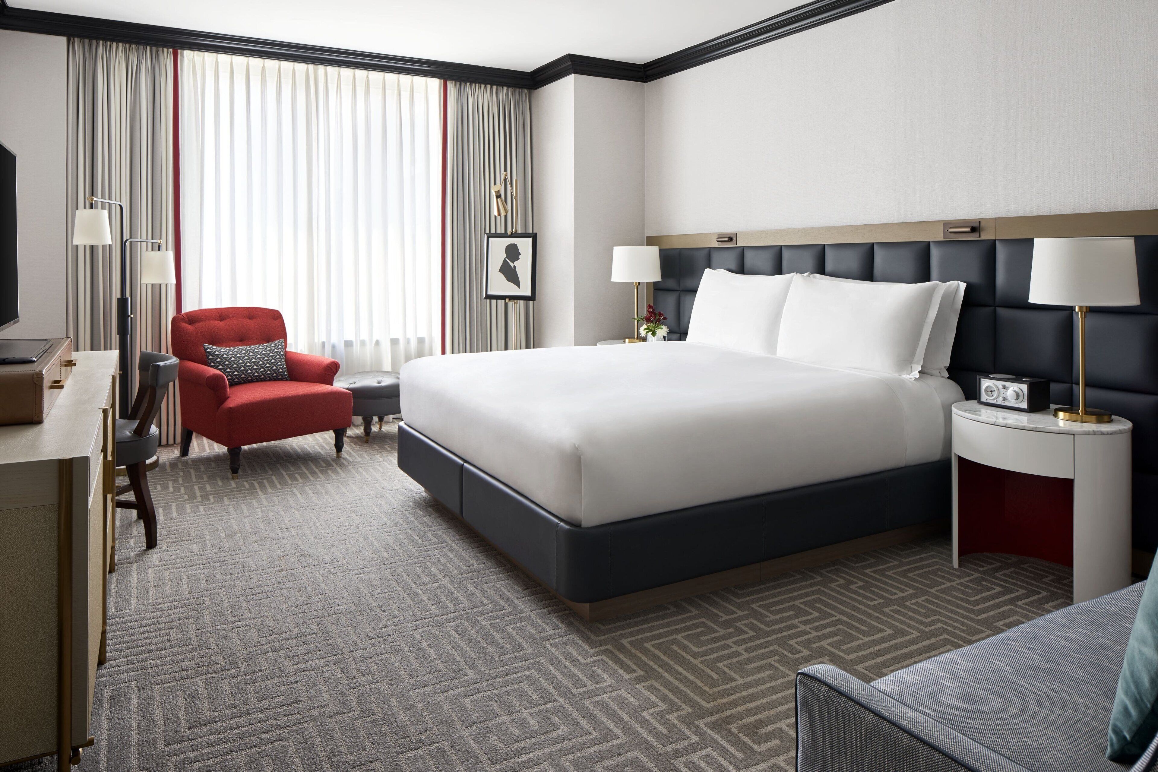 A hotel room with a large bed, a red armchair, a desk with a lamp, and a TV.