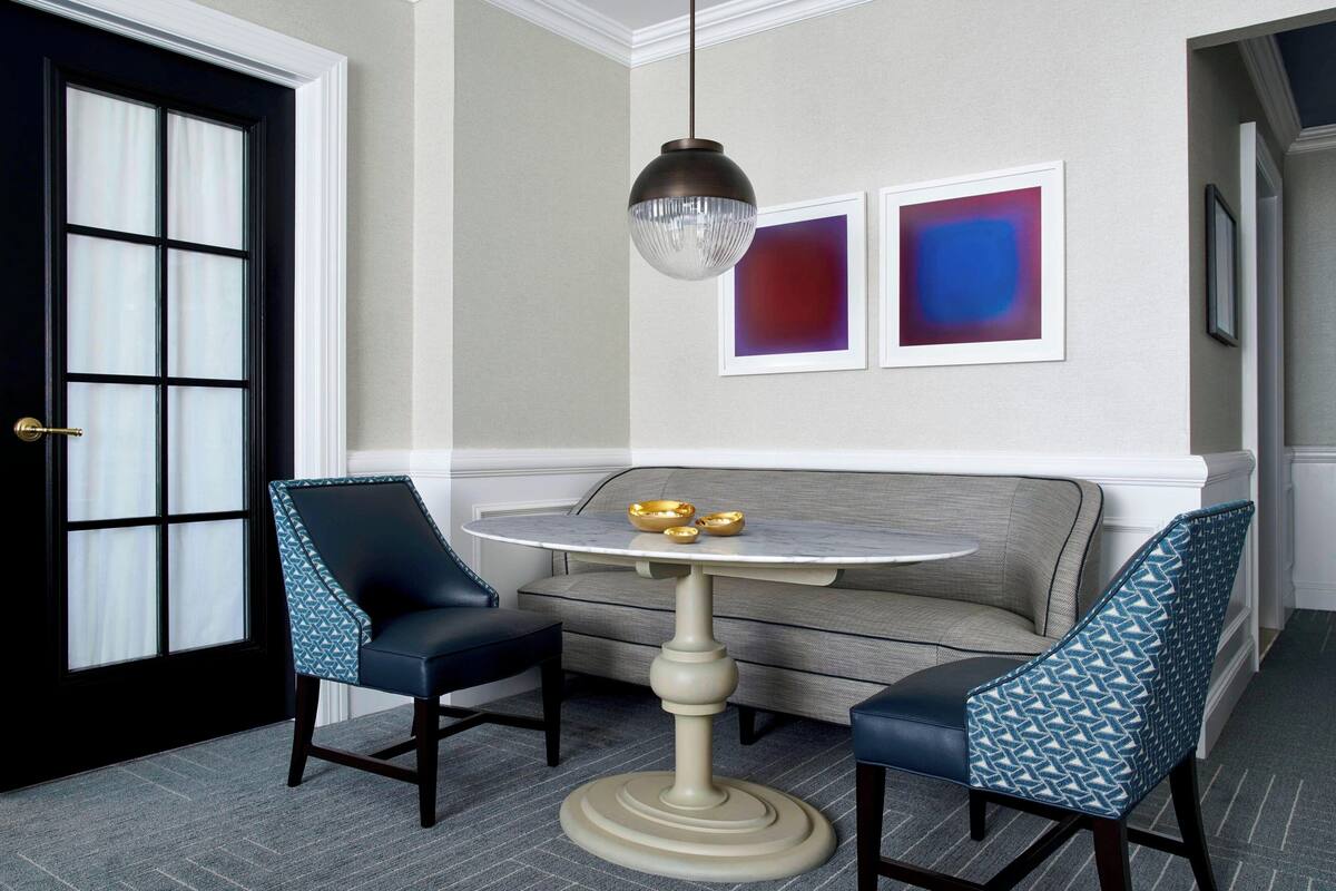 A room with a round table, two chairs, a sofa, and two abstract paintings on the wall.