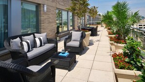 Un patio exterior con mobiliario de mimbre, macetas con flores y un edificio de ladrillo al fondo.