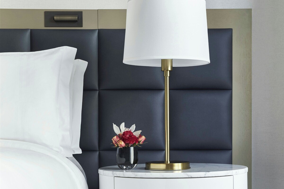 A neatly made bed with white linens, a bedside table with a vase of flowers, and a modern lamp with a gold base.
